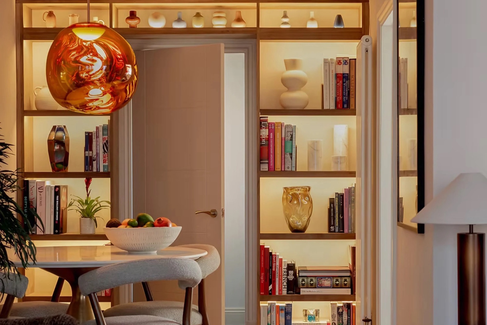Warmly lit living and dining room with oak shelving, books and ceramics